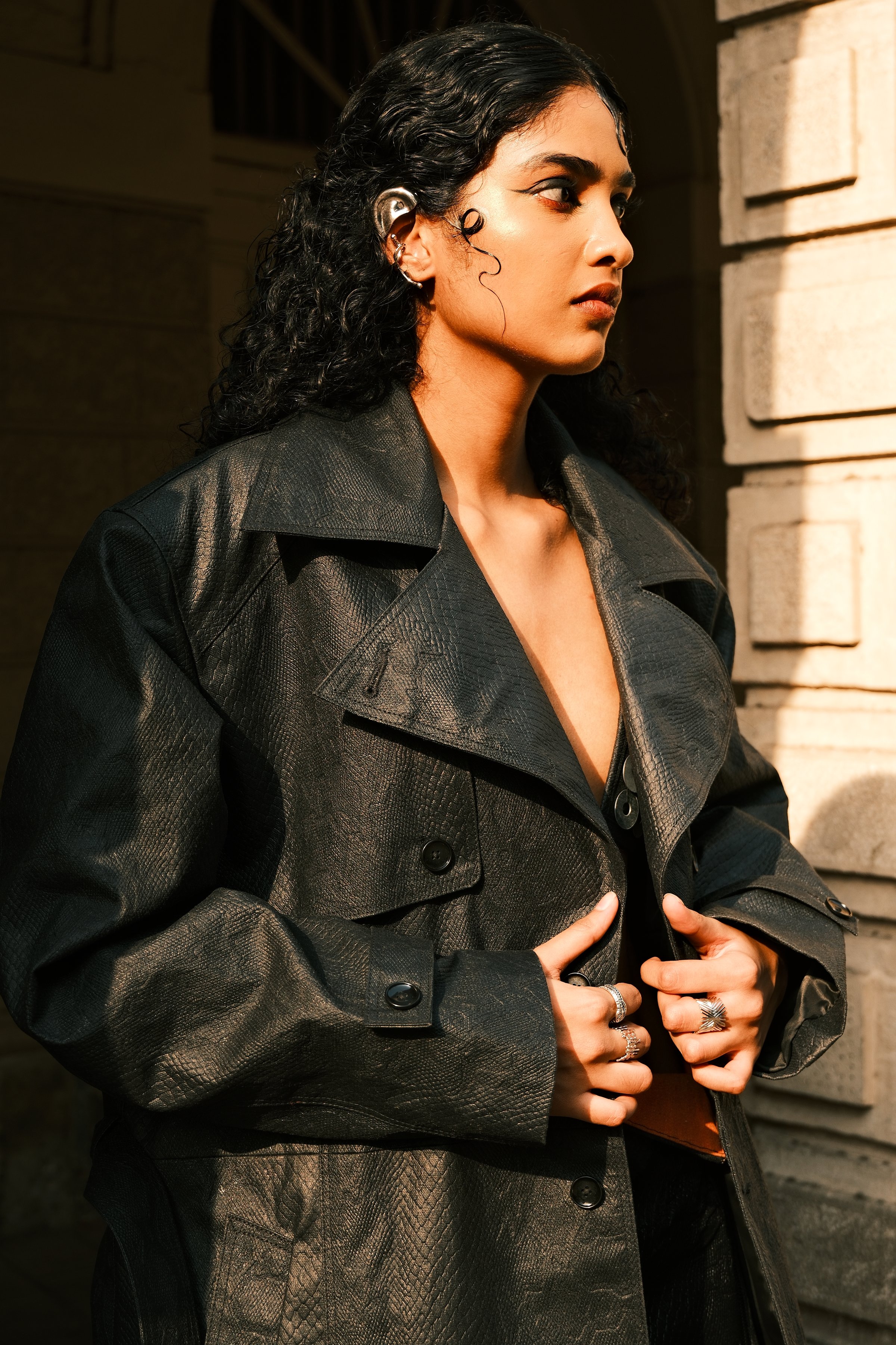 A model with curly hair in a leather trench against a pillar