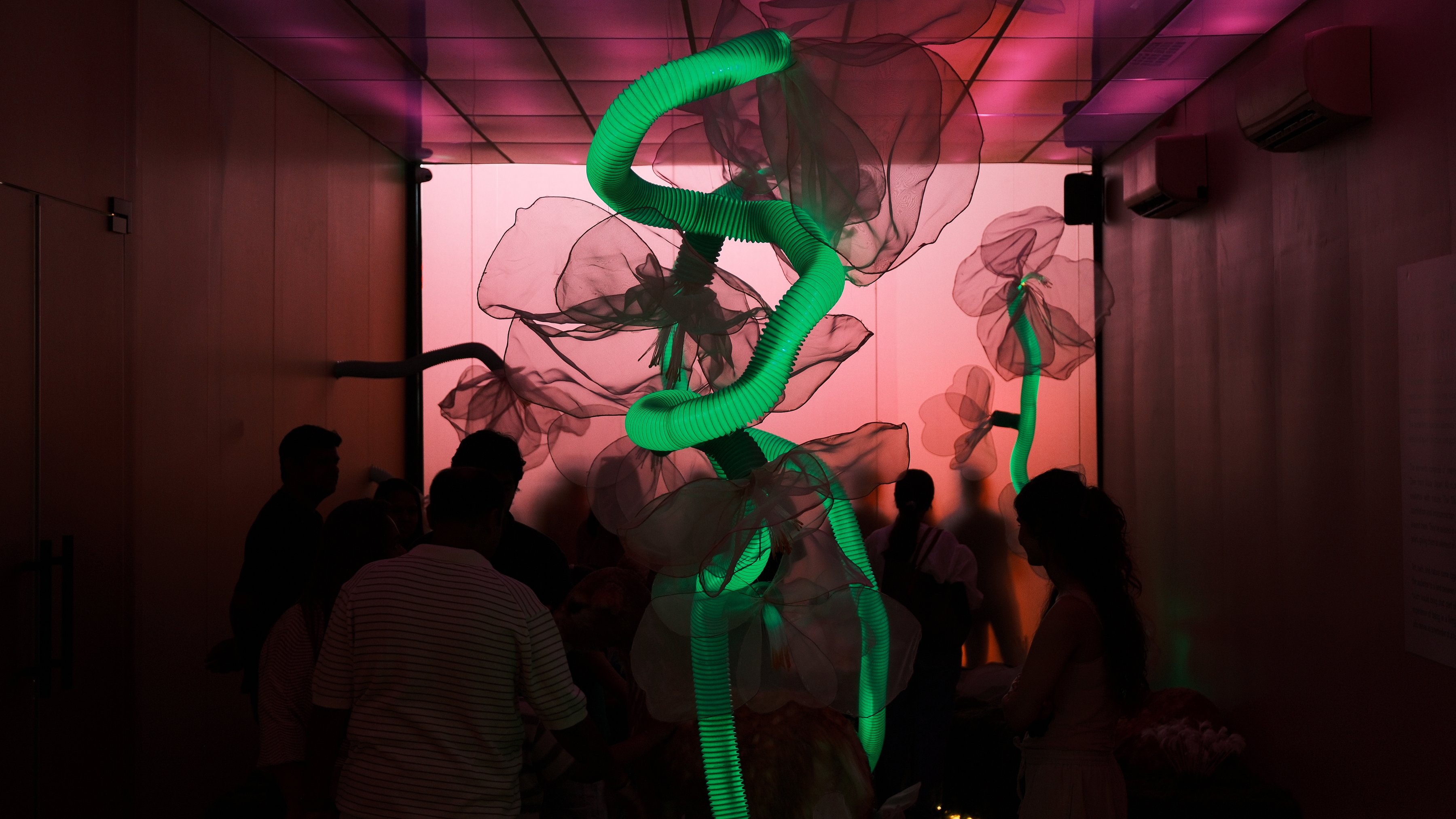 A green neon sculpture among sheer glass flowers in a dark room full of visitors