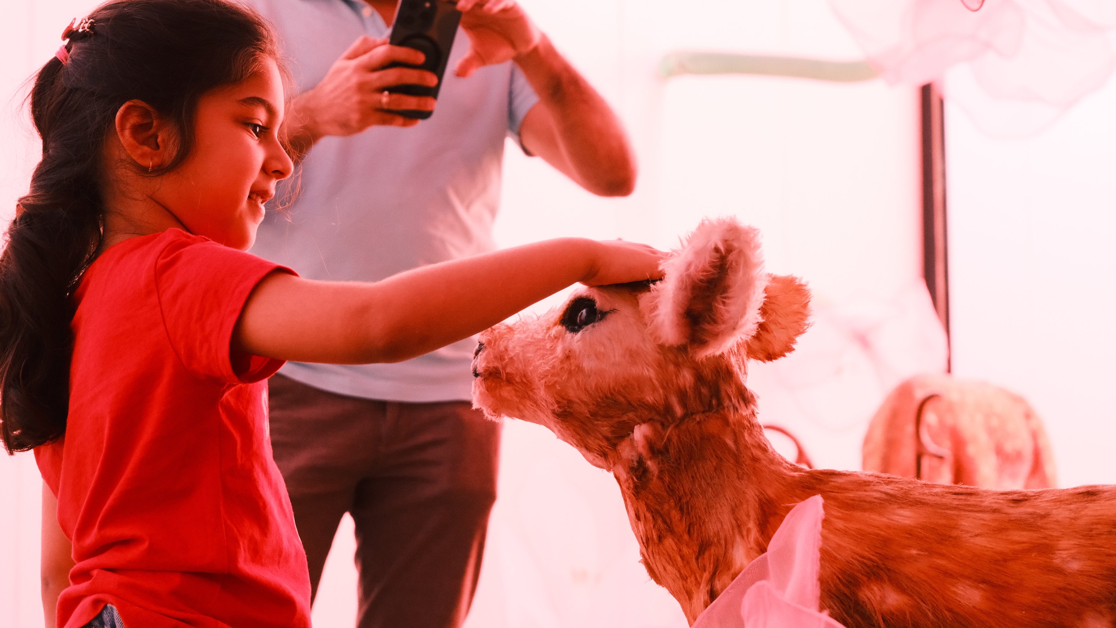 A girl in red reaching out to a fawn as her father takes a photo