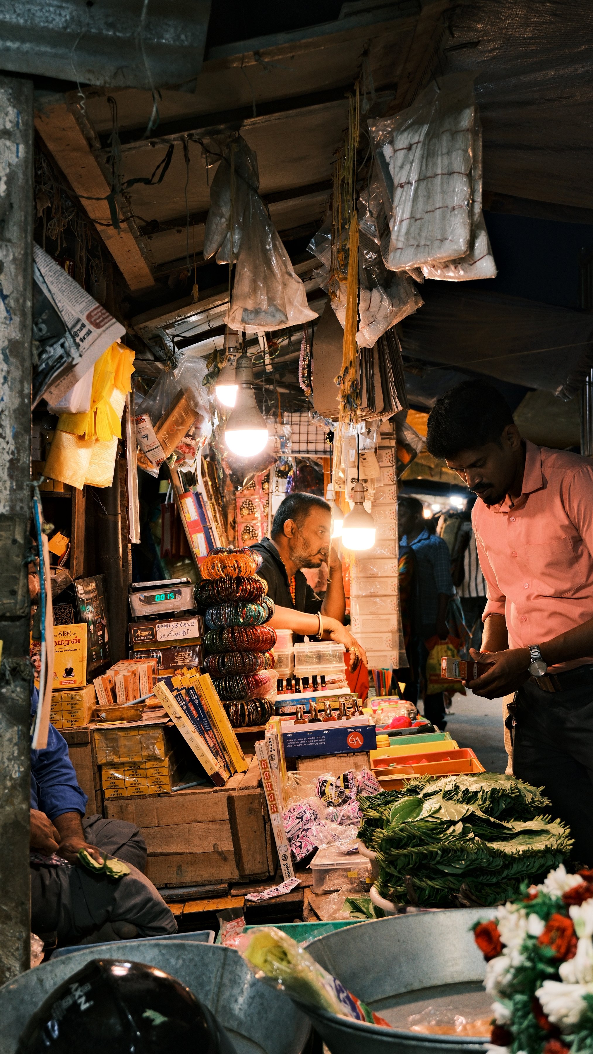 A market stall at night lit by bare bulbs, piled with goods
