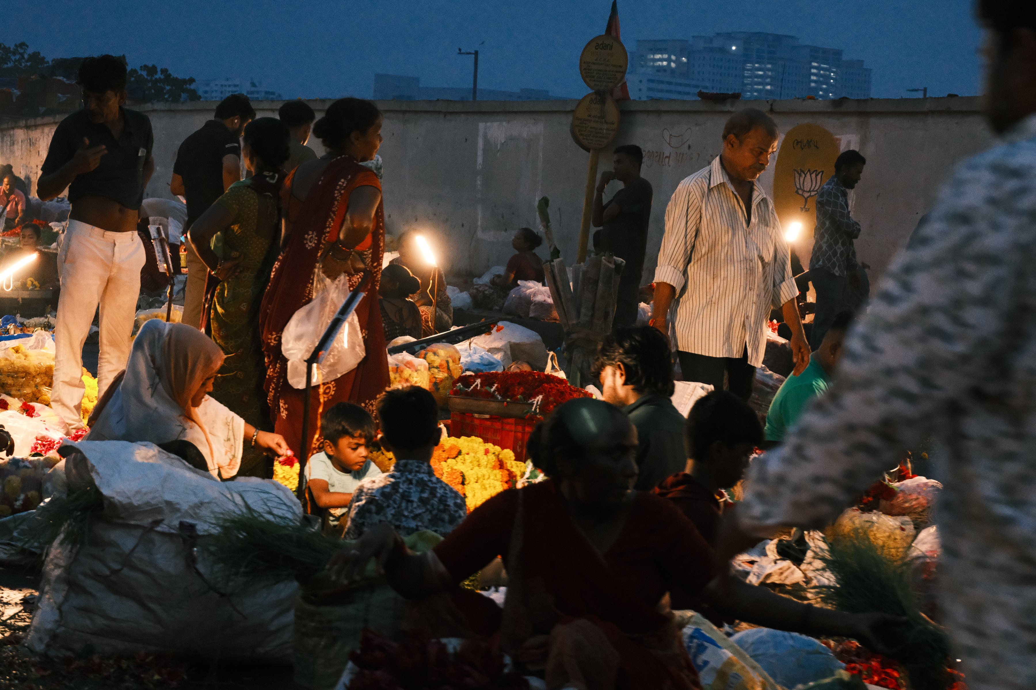 A wide view of the market floor, sellers crouched among sacks of flowers
