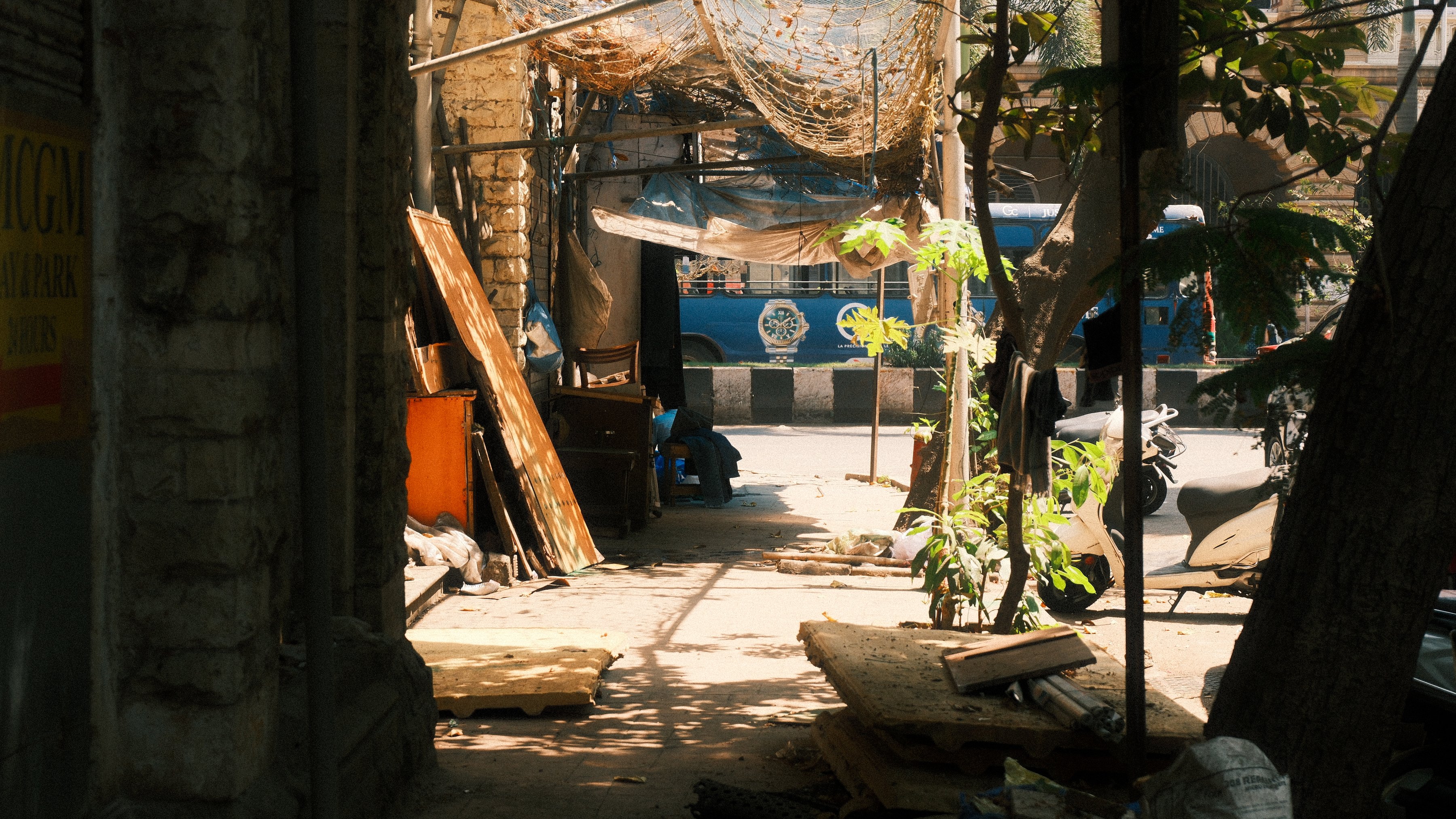 A shaded side street opening onto a sunlit road and a passing blue bus