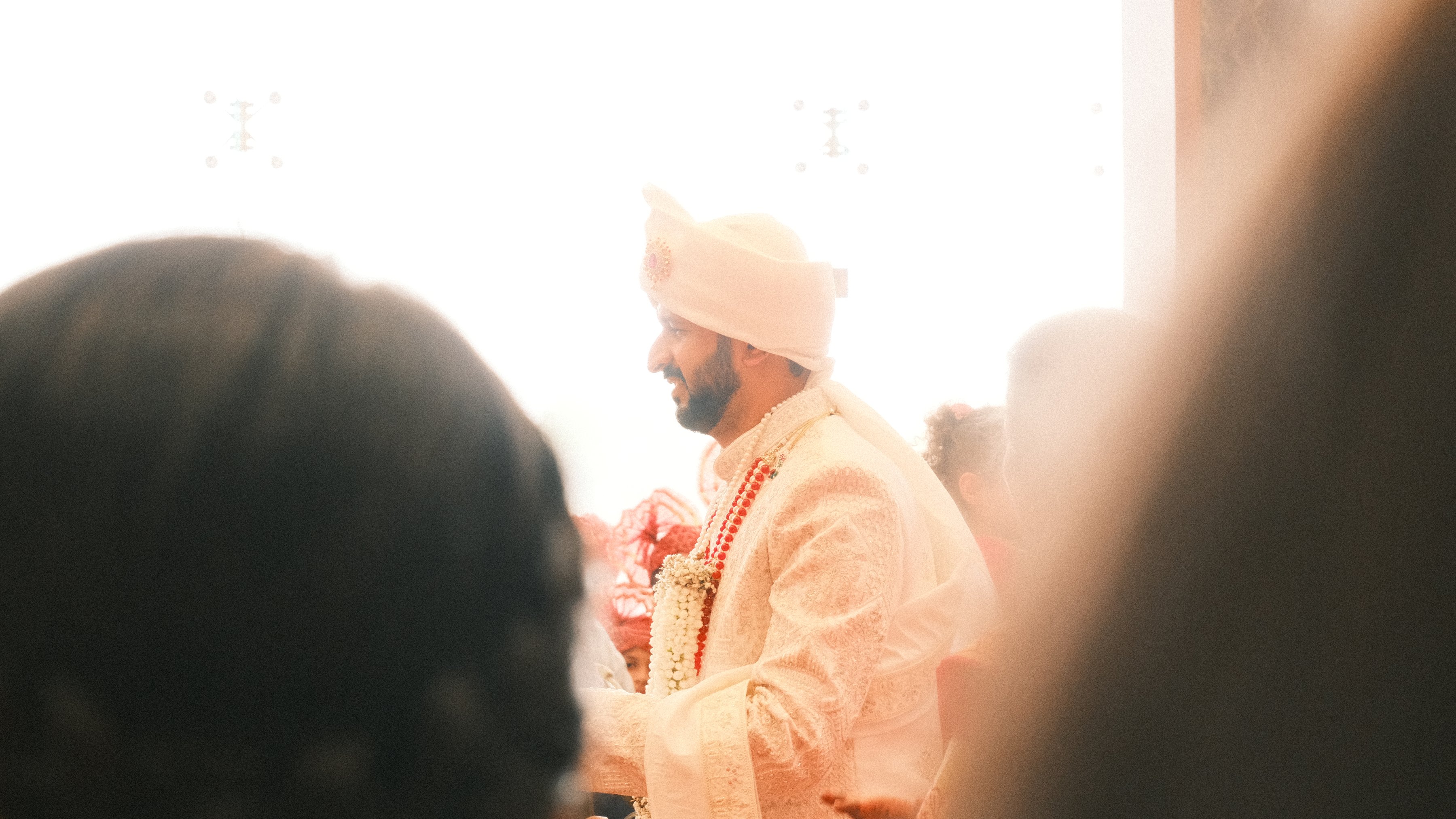 The groom smiling in a cream sherwani, framed by guests