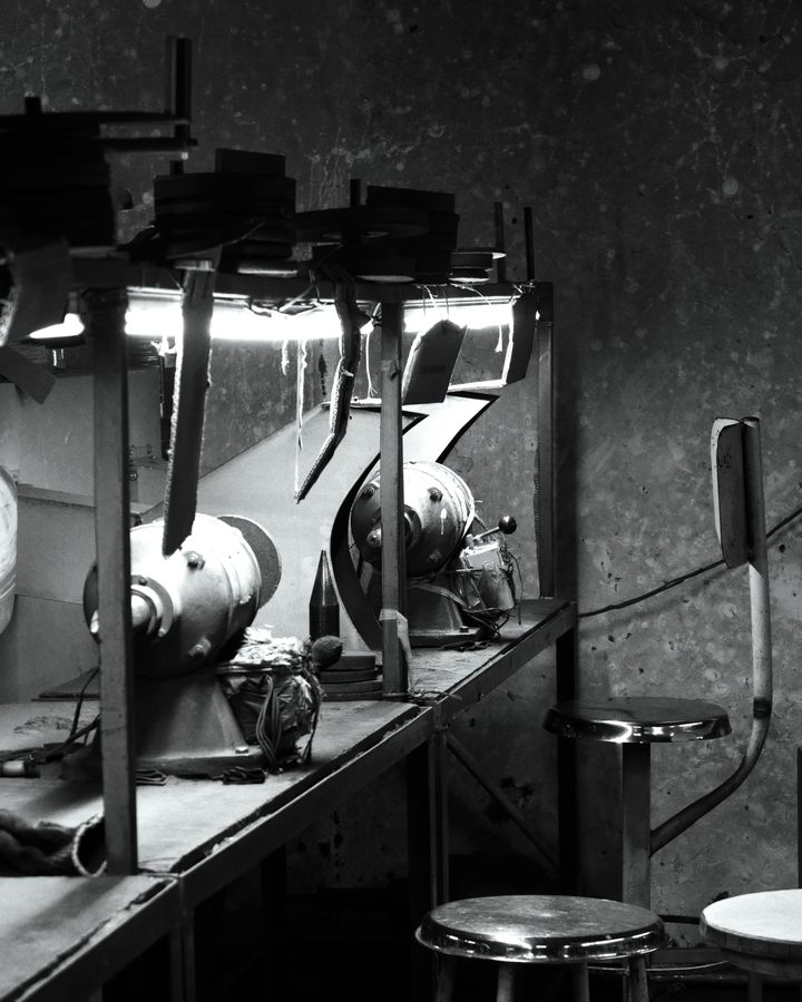A row of grinding stations and stools under bright lamps on the factory floor, in black and white