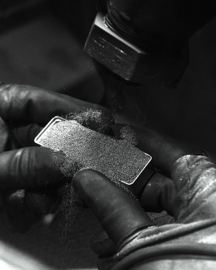 Gloved hands sanding a small metal plate as dust flies from the grinder, in black and white