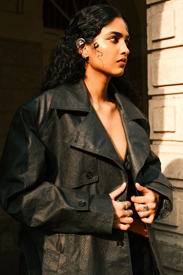 A model with curly hair in a leather trench against a pillar