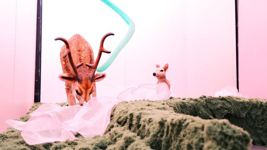 A deer sculpture grazing on a mossy mound in a pink room, a fawn behind it