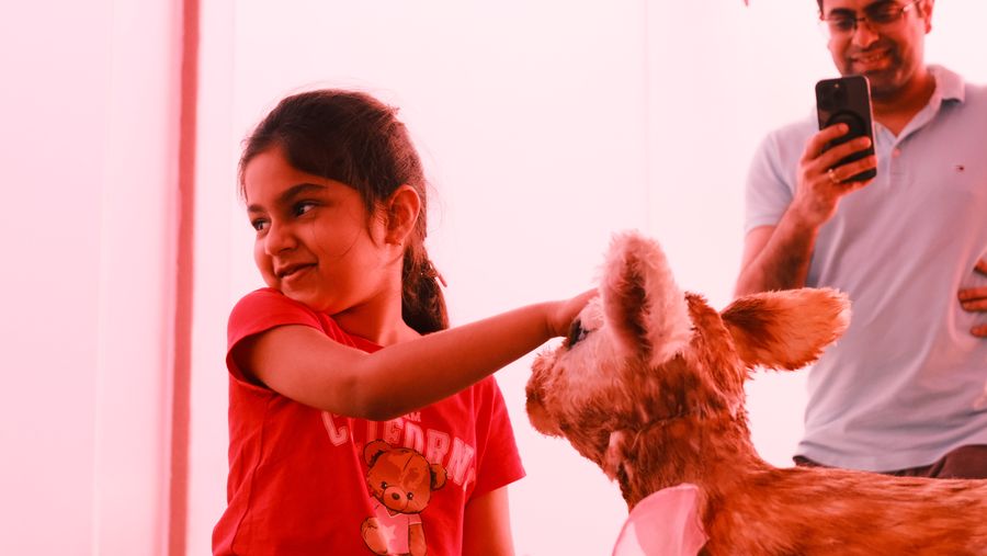 A girl in red smiling as she pets a fawn, her father filming behind her