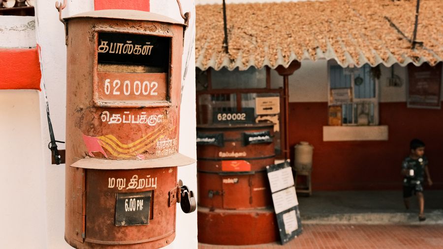 A rusted red post box marked 620002 outside a small post office, a boy in the doorway