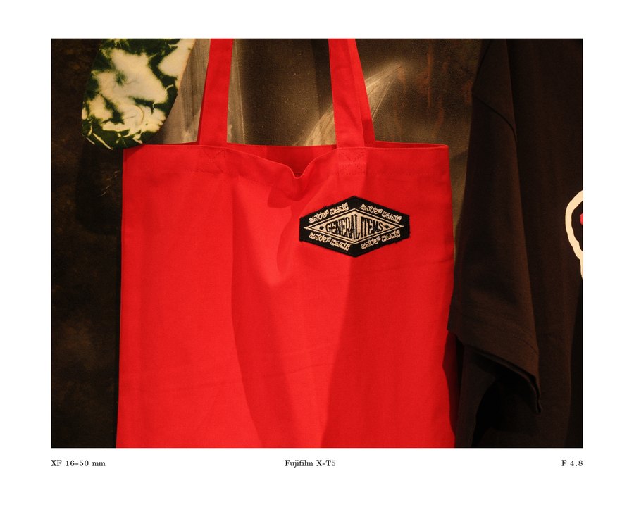 A red General Items tote bag hanging against a dark wall, presented with a printed caption