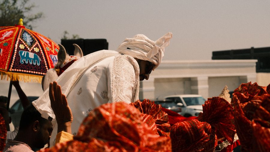The groom in a white turban bowing under a bright umbrella among red turbans